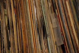 A close-up view of a large collection of vinyl records, tightly packed together in an upright position. The edges of the record sleeves are visible, showcasing a variety of colors, textures, and print styles. The records are stacked vertically, creating a layered effect with a mix of faded and vibrant hues.