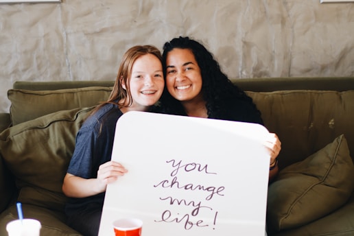 Two people are sitting on a green couch, smiling and holding a white sign with the handwritten message 'you change my life!'. A red cup is visible on the foreground.