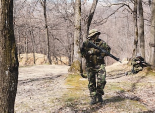 In a forested area with bare trees, two individuals dressed in camouflage uniforms are holding rifles. One person is standing and walking cautiously, while the other is kneeling and aiming their rifle.