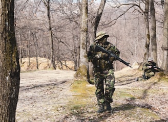 In a forested area with bare trees, two individuals dressed in camouflage uniforms are holding rifles. One person is standing and walking cautiously, while the other is kneeling and aiming their rifle.
