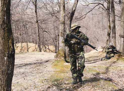 In a forested area with bare trees, two individuals dressed in camouflage uniforms are holding rifles. One person is standing and walking cautiously, while the other is kneeling and aiming their rifle.