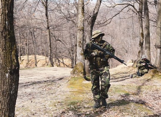 In a forested area with bare trees, two individuals dressed in camouflage uniforms are holding rifles. One person is standing and walking cautiously, while the other is kneeling and aiming their rifle.