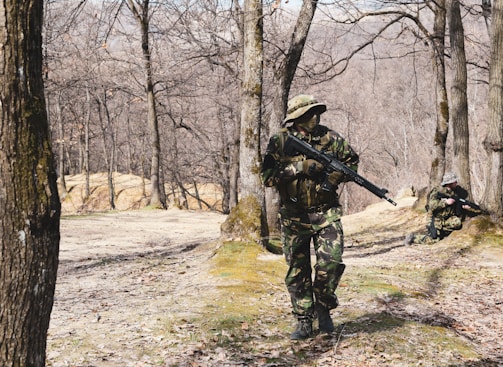 In a forested area with bare trees, two individuals dressed in camouflage uniforms are holding rifles. One person is standing and walking cautiously, while the other is kneeling and aiming their rifle.