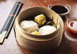 A wooden bamboo steamer basket filled with dim sum, including translucent dumplings and siu mai. Next to it are two small bowls of dipping sauces and a pair of black chopsticks with red accents, all on a wooden table.