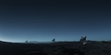 Three large satellite dishes stand on a dark, barren landscape under a deep blue sky. The scene appears to be desolate, with rocky terrain extending into the distance. The satellite dishes are evenly spaced and pointed towards the sky, suggesting a purpose of capturing signals from space. The horizon line is barely visible, shrouded in the twilight or early dawn light, creating a serene and somewhat mysterious atmosphere.