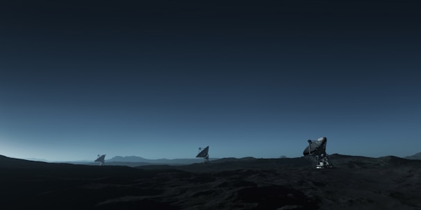 Three large satellite dishes stand on a dark, barren landscape under a deep blue sky. The scene appears to be desolate, with rocky terrain extending into the distance. The satellite dishes are evenly spaced and pointed towards the sky, suggesting a purpose of capturing signals from space. The horizon line is barely visible, shrouded in the twilight or early dawn light, creating a serene and somewhat mysterious atmosphere.