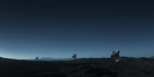 Three large satellite dishes stand on a dark, barren landscape under a deep blue sky. The scene appears to be desolate, with rocky terrain extending into the distance. The satellite dishes are evenly spaced and pointed towards the sky, suggesting a purpose of capturing signals from space. The horizon line is barely visible, shrouded in the twilight or early dawn light, creating a serene and somewhat mysterious atmosphere.