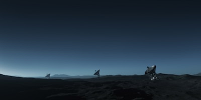 Three large satellite dishes stand on a dark, barren landscape under a deep blue sky. The scene appears to be desolate, with rocky terrain extending into the distance. The satellite dishes are evenly spaced and pointed towards the sky, suggesting a purpose of capturing signals from space. The horizon line is barely visible, shrouded in the twilight or early dawn light, creating a serene and somewhat mysterious atmosphere.