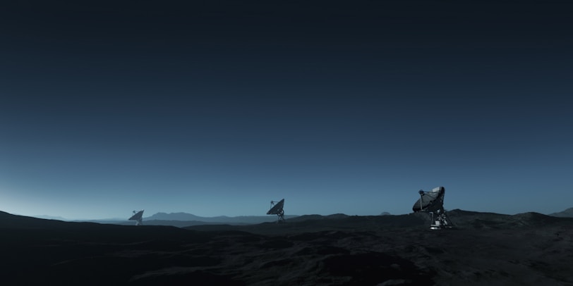 Three large satellite dishes stand on a dark, barren landscape under a deep blue sky. The scene appears to be desolate, with rocky terrain extending into the distance. The satellite dishes are evenly spaced and pointed towards the sky, suggesting a purpose of capturing signals from space. The horizon line is barely visible, shrouded in the twilight or early dawn light, creating a serene and somewhat mysterious atmosphere.