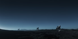 Three large satellite dishes stand on a dark, barren landscape under a deep blue sky. The scene appears to be desolate, with rocky terrain extending into the distance. The satellite dishes are evenly spaced and pointed towards the sky, suggesting a purpose of capturing signals from space. The horizon line is barely visible, shrouded in the twilight or early dawn light, creating a serene and somewhat mysterious atmosphere.