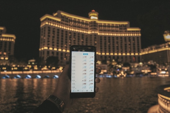 A hand holding a smartphone displaying a financial or trading app, with a large, well-lit hotel building in the background. The building features rows of illuminated windows and the scene is set at night, reflected in the water in front.