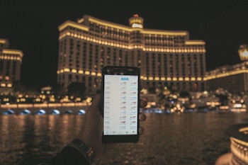 A hand holding a smartphone displaying a financial or trading app, with a large, well-lit hotel building in the background. The building features rows of illuminated windows and the scene is set at night, reflected in the water in front.