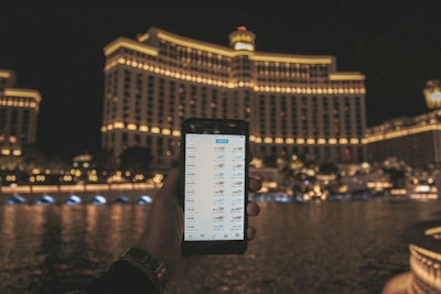A hand holding a smartphone displaying a financial or trading app, with a large, well-lit hotel building in the background. The building features rows of illuminated windows and the scene is set at night, reflected in the water in front.