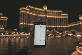 A hand holding a smartphone displaying a financial or trading app, with a large, well-lit hotel building in the background. The building features rows of illuminated windows and the scene is set at night, reflected in the water in front.