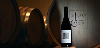 A bottle of wine stands in front of large wooden barrels. The barrels are marked with the logo 'la JASSE CASTEL.' The lighting is warm, focusing on the bottle, which has a prominent label displaying 'Les Combarelles 2017.'