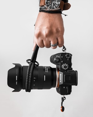 A hand holding a Sony camera with a large Tamron lens attached. The person is wearing a watch with a brown strap and a black braided bracelet. The background is a simple, light grey color.