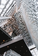 A futuristic interior featuring a complex geometric ceiling design made up of hexagonal glass and metal structures, creating a lattice-like pattern. The space is illuminated with natural light, showcasing sleek, modern architectural elements and dark shaded staircases or walkways that contrast with the bright ceiling.