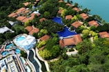An aerial view of a resort with several red-roofed villas nestled among lush greenery. A large, winding swimming pool runs through the complex. There is also a water park section featuring multiple pools and slides. The resort is located near a body of water, possibly a lake or sea.