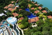 An aerial view of a resort with several red-roofed villas nestled among lush greenery. A large, winding swimming pool runs through the complex. There is also a water park section featuring multiple pools and slides. The resort is located near a body of water, possibly a lake or sea.