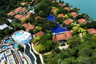 An aerial view of a resort with several red-roofed villas nestled among lush greenery. A large, winding swimming pool runs through the complex. There is also a water park section featuring multiple pools and slides. The resort is located near a body of water, possibly a lake or sea.