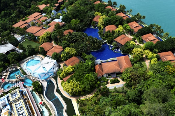 An aerial view of a resort with several red-roofed villas nestled among lush greenery. A large, winding swimming pool runs through the complex. There is also a water park section featuring multiple pools and slides. The resort is located near a body of water, possibly a lake or sea.
