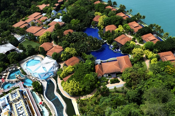 An aerial view of a resort with several red-roofed villas nestled among lush greenery. A large, winding swimming pool runs through the complex. There is also a water park section featuring multiple pools and slides. The resort is located near a body of water, possibly a lake or sea.