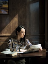 A woman with long dark hair sits at a wooden table in a warmly lit room. She is reading a newspaper titled 'The Astana Times' and has a focused expression. On the table, there is an open book and a white cup. The wall behind her is made of wooden panels, adding to the cozy atmosphere.