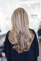Long, wavy hair with blonde and light brown highlights cascading down the back of a person wearing a dark top. The background is a blurred indoor setting, suggesting a salon environment.