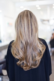 Long, wavy hair with blonde and light brown highlights cascading down the back of a person wearing a dark top. The background is a blurred indoor setting, suggesting a salon environment.