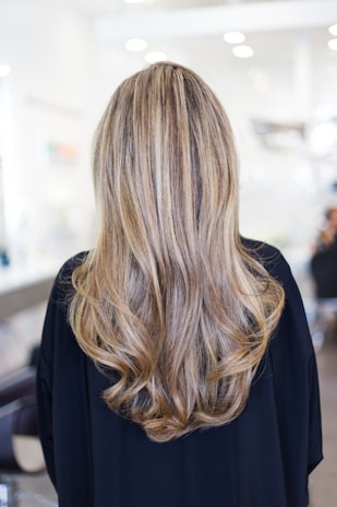 Long, wavy hair with blonde and light brown highlights cascading down the back of a person wearing a dark top. The background is a blurred indoor setting, suggesting a salon environment.