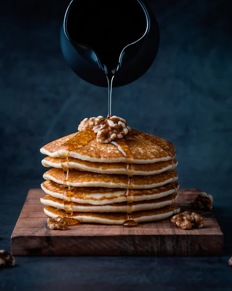A short stack of mini pancakes topped with banana slices and a yoghurt pot on a pale plate