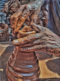 A pair of hands covered in clay skillfully shaping a piece of pottery on a spinning wheel. The artist's movements appear focused and precise, with textured details of the clay and hands prominently visible against a background of earthy tones.