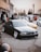 A black, modified BMW is parked in a lot, surrounded by other cars and a group of people. The car has lowered suspension and shiny chrome wheels. In the background, there are trees and a building with signage, and a few people standing and conversing.