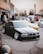 A black, modified BMW is parked in a lot, surrounded by other cars and a group of people. The car has lowered suspension and shiny chrome wheels. In the background, there are trees and a building with signage, and a few people standing and conversing.