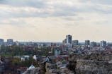 A cityscape featuring a mix of modern high-rise buildings and older architecture spread across a densely populated area. In the foreground, there are ancient stone ruins, where a person is sitting on the rocks, enjoying the view. Trees and green spaces are interspersed between the buildings, and a flock of birds is flying in the cloudy sky.