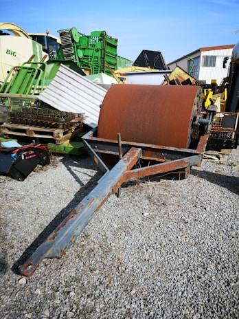 A large, rusted metal drum is placed on a gravel surface, surrounded by various pieces of agricultural machinery. The background includes several green and yellow equipments, some stacked or leaning against each other, suggesting a storage or scrapyard setting.