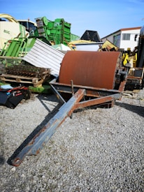 A large, rusted metal drum is placed on a gravel surface, surrounded by various pieces of agricultural machinery. The background includes several green and yellow equipments, some stacked or leaning against each other, suggesting a storage or scrapyard setting.
