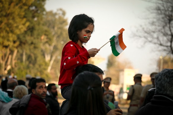 A young child wearing a red sweater with heart patterns is sitting on someone’s shoulders in a crowd. The child is holding a small Indian flag and has painted it on their face. In the background, trees and other people, possibly in uniform, can be seen slightly out of focus.