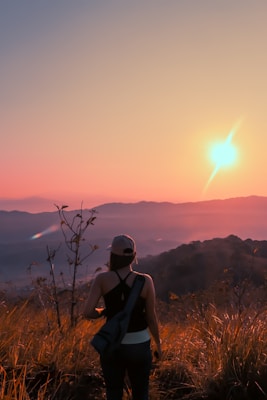 A person stands on a hilltop, gazing at a vibrant sunset over distant mountains. The sky is painted in shades of orange and pink, casting a warm glow over the landscape. The person wears a backpack and cap, surrounded by tall grass swaying gently in the evening breeze.