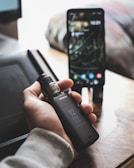A person is holding a vaporesso vape mod in their hand. In the background, there is a smartphone placed on a wooden surface, displaying a lock screen. The setting appears to be indoors, possibly a home or office environment, with soft natural light illuminating the scene.