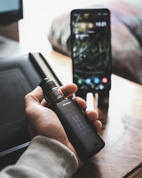 A person is holding a vaporesso vape mod in their hand. In the background, there is a smartphone placed on a wooden surface, displaying a lock screen. The setting appears to be indoors, possibly a home or office environment, with soft natural light illuminating the scene.