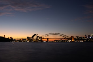 A serene view of a cityscape at dusk featuring the iconic Sydney Opera House with its distinct sails and the Sydney Harbour Bridge illuminated against the darkening sky. The skyline is lit up with city lights, reflecting a calm and peaceful atmosphere as daylight fades.