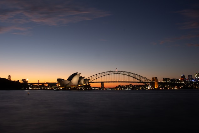 A serene view of a cityscape at dusk featuring the iconic Sydney Opera House with its distinct sails and the Sydney Harbour Bridge illuminated against the darkening sky. The skyline is lit up with city lights, reflecting a calm and peaceful atmosphere as daylight fades.