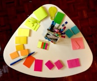 A white triangular table is covered with colorful stationery items. Various notepads and sticky notes in neon yellow, green, blue, pink, and orange are arranged in a semi-circle. A clear container holds multiple colored markers, pens, and highlighters. A blue pen is also visible on the table alongside some multicolored sticky tabs.