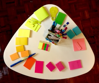 A white triangular table is covered with colorful stationery items. Various notepads and sticky notes in neon yellow, green, blue, pink, and orange are arranged in a semi-circle. A clear container holds multiple colored markers, pens, and highlighters. A blue pen is also visible on the table alongside some multicolored sticky tabs.