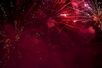 Explosions of vibrant fireworks light up the night sky with bursts of red and sparkling white trails. Smoky clouds create a dramatic backdrop, enhancing the visual spectacle.