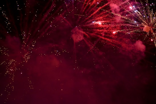 Explosions of vibrant fireworks light up the night sky with bursts of red and sparkling white trails. Smoky clouds create a dramatic backdrop, enhancing the visual spectacle.