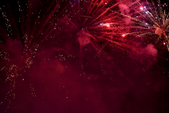 Explosions of vibrant fireworks light up the night sky with bursts of red and sparkling white trails. Smoky clouds create a dramatic backdrop, enhancing the visual spectacle.