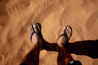 A pair of feet in black flip-flops standing on sunlit, reddish-brown sand. The feet are positioned slightly apart, with shadows cast on the sand surface. The sand is textured with small pebbles and fine grains.