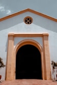 A facade of a historic building with white walls and a large wooden doorway framed by a light brown arch. Above the doorway, there is a small circular window with a decorative design. The building has a simple, symmetrical architecture with soft shadows cast on its surface.